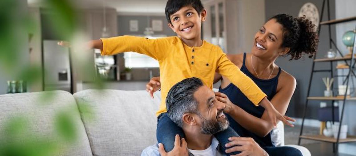 Cheerful indian son sitting on father shoulder playing at home with african mother. Playful little boy enjoying spending time with parents at home. Flying child enjoying playing with his ethnic family